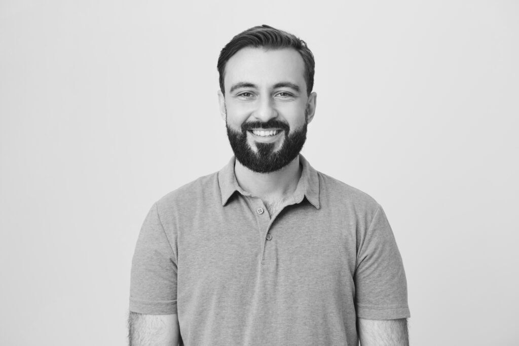 Good-looking european male adult with beard and moustache smiling broadly over gray background. Boyfriend stand behind door, knocks and waits till girlfriend will open it